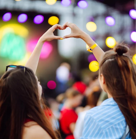 Twee vrienden vormen een hartje met hun handen terwijl ze naar een live muziekoptreden kijken op een openluchtfestival met kleurrijke podiumverlichting.