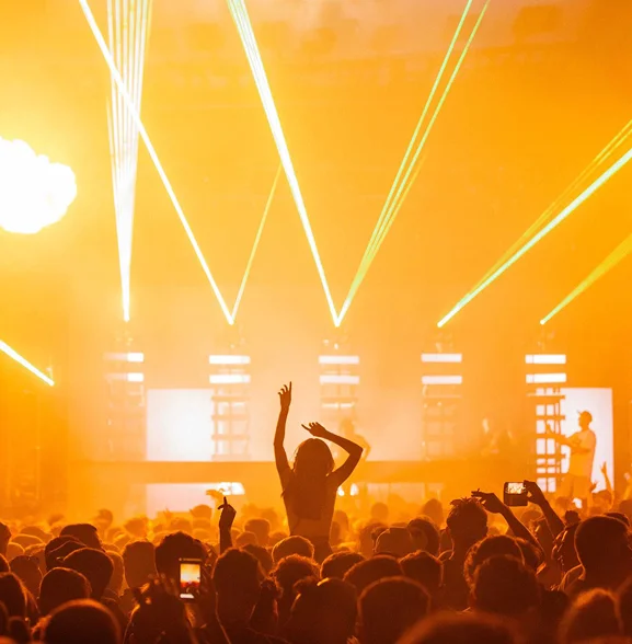 Grote menigte danst op een live muziekfestival met feloranje podiumverlichting, lasers en een DJ die op het podium optreedt.