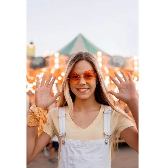 Lachende jonge vrouw met oranje zonnebril poseert op een kermis of festival met carrouselverlichting op de achtergrond.